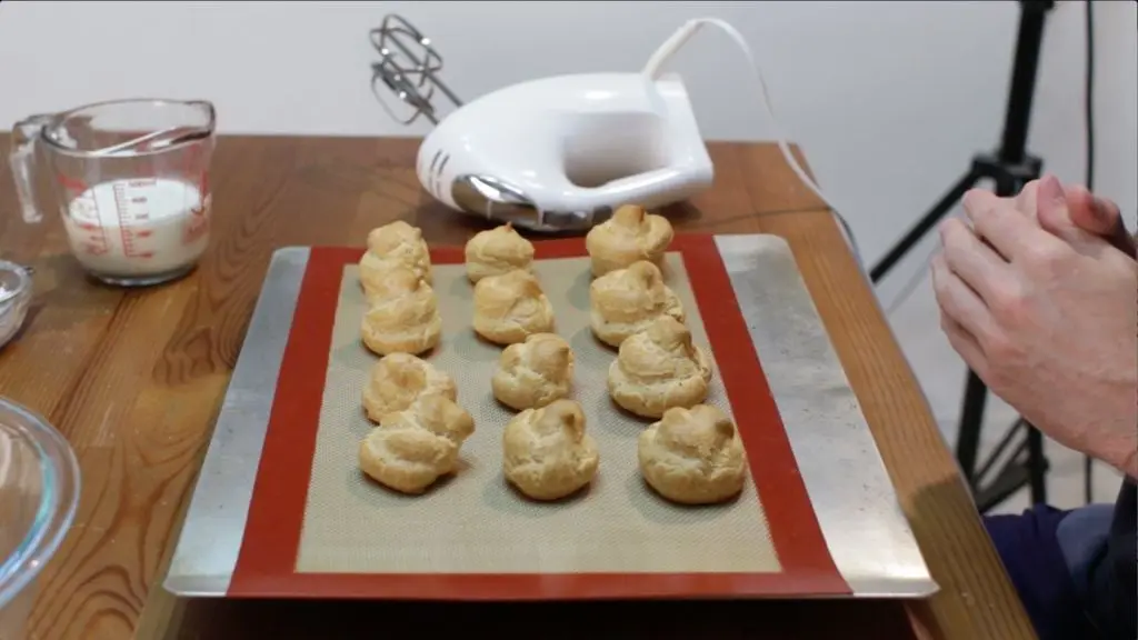 Freshly baked cream puffs resting on a silicone-mat lined cookie sheet on a table. 