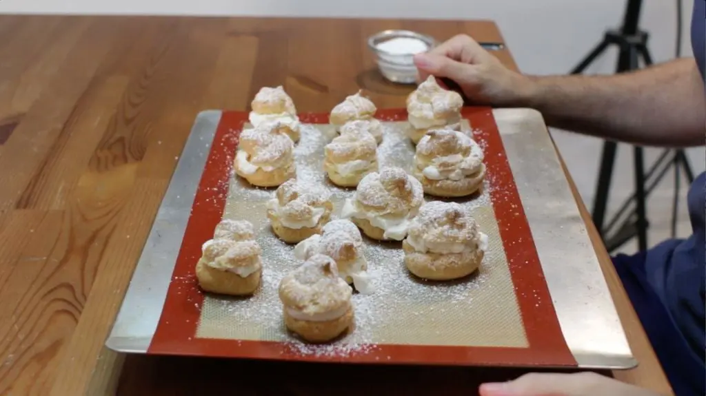 12 finished cream puffs topped with powder sugar on a silicone mat lined cookie pan.