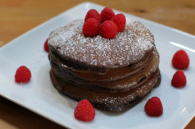 Chocolate pancakes stacked on a white plate covered in powdered sugar and raspberries