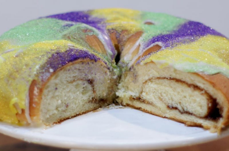 Mardi Gras king cake on a white cake pedestal on a wooden table