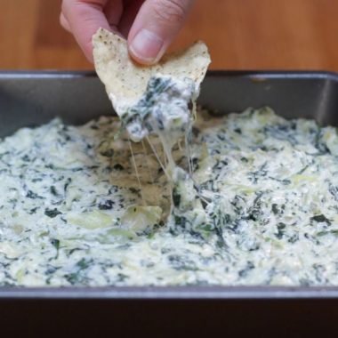 Spinach Artichoke dip in a square metal pan and hand dipping a chip in it.
