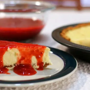 Easy cheesecake recipe with a slice of cheesecake covered in strawberry sauce on a table.