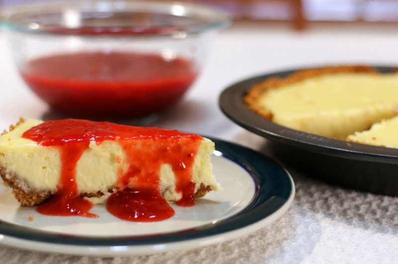 Easy cheesecake recipe with a slice of cheesecake covered in strawberry sauce on a table.