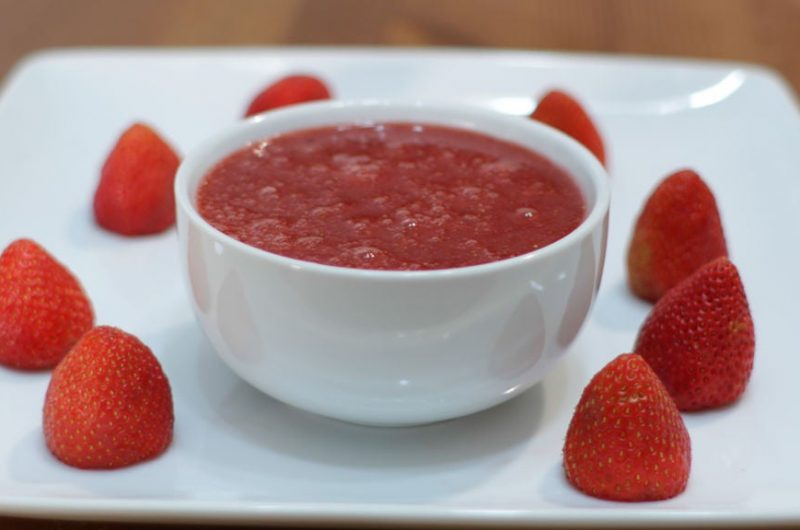 Homemade strawberry sauce in a white bowl on a plate surrounded by whole strawberries