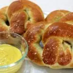 Three homemade soft pretzels on a plate next to a bowl of mustard.