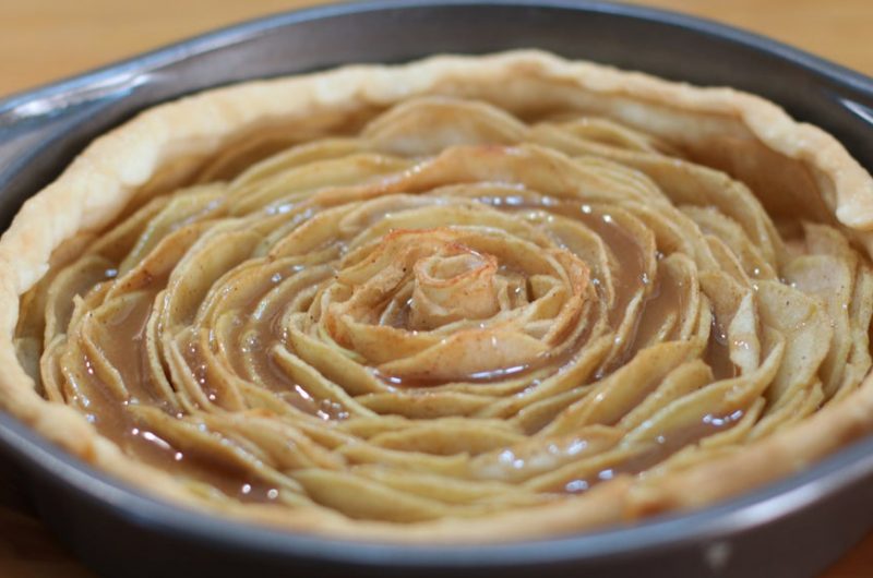 Caramel rose apple pie in a metal pie pan on a wooden table.