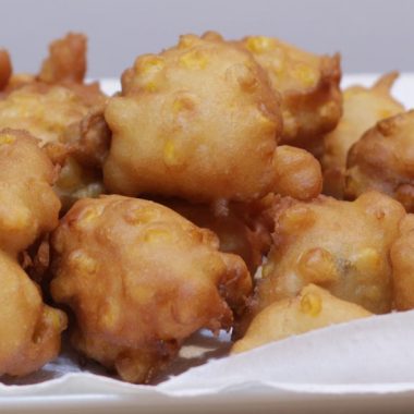 Homemade corn fritters pile up on a plate with paper towel.