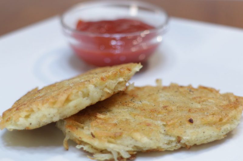 Two crispy homemade hash browns on a white plate next to bowl of ketchup