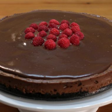 Triple chocolate cheesecake with raspberries on top of a white cake pedestal.