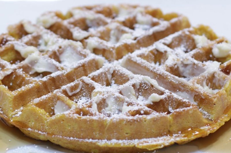 Pumpkin waffles on a white plate with butter, maple syrup, and powdered sugar.
