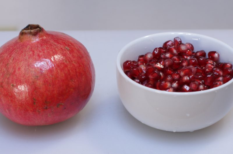 a pomegranate on a white cutting board next to a bowl of pomegranate seeds