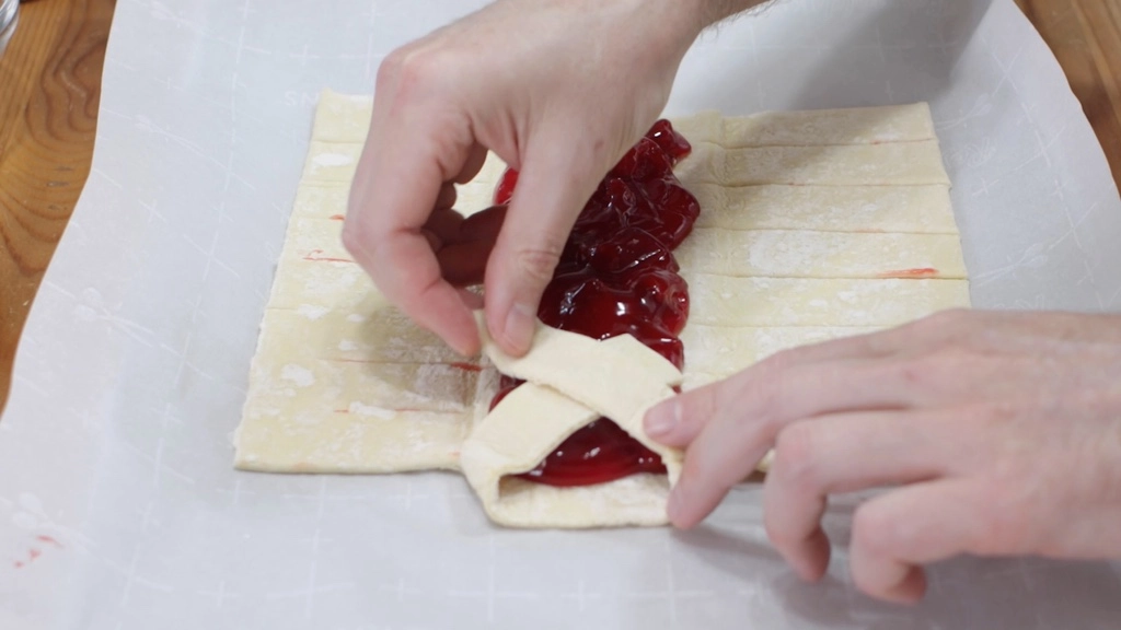 Fingers folding strips of puff pastry over the middle of the strawberry filling.