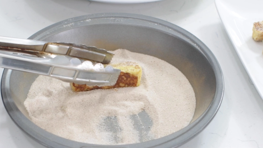Tongs rolling cinnamon French toast sticks in cinnamon and sugar.