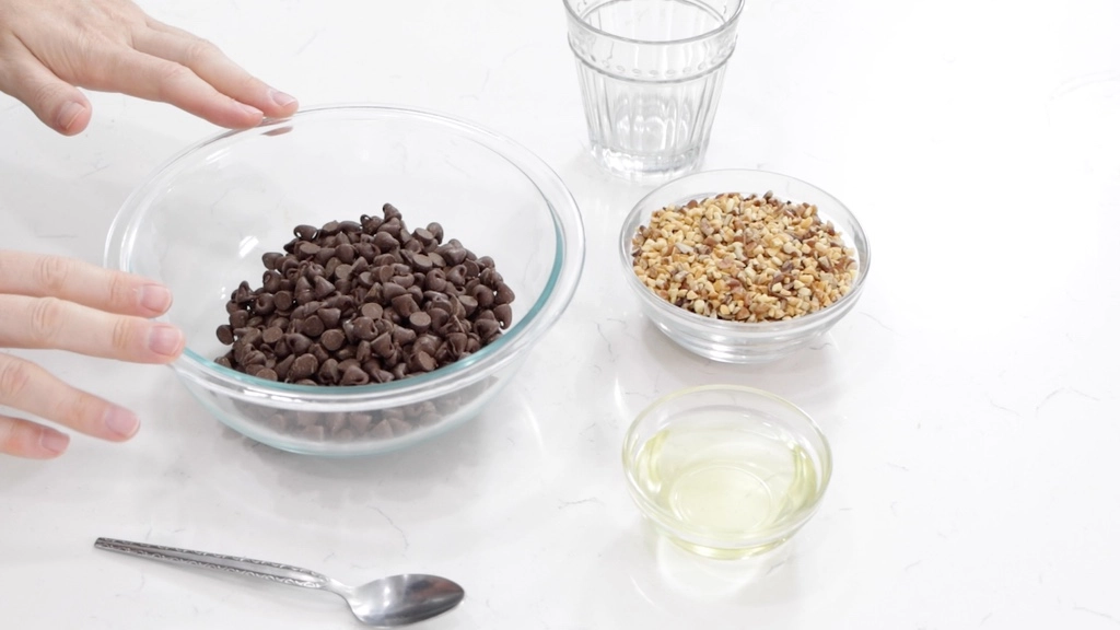 Ice Cream Bars Recipe | 5 Ingredients - In The Kitchen With Matt 5 Hands resting on a glass bowl full of chocolate chips next to a bowl of nuts.