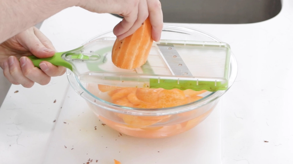Hand slicing a peeled sweet potato on a mandolin.
