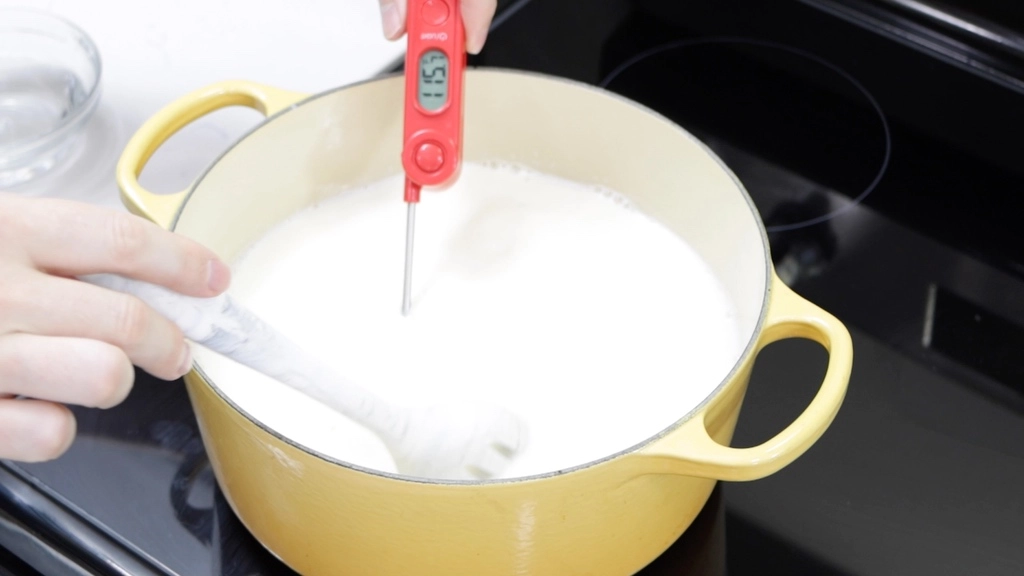 Hand with red thermometer in milk on a stovetop.