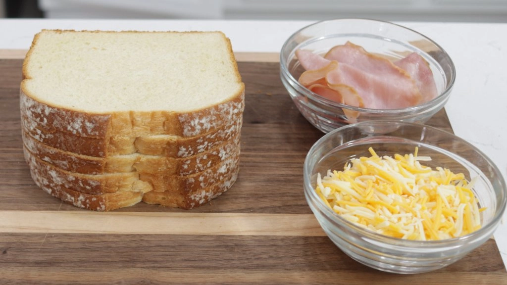 Slices of white bread next to a bowl of ham and cheese on a wooden cutting board.