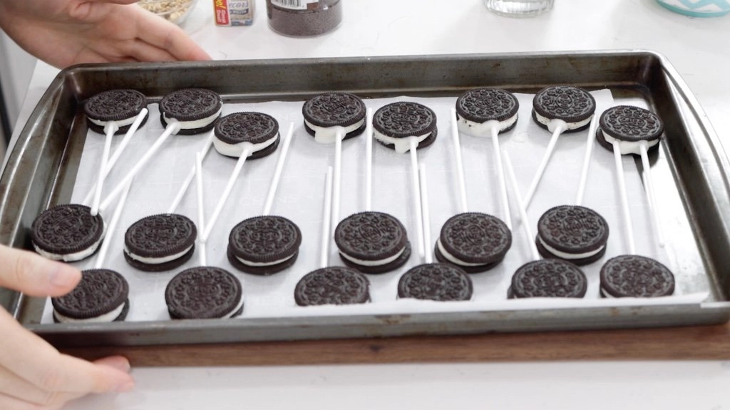 Several Oreos with sticks in them on a sheet pan with parchment paper.