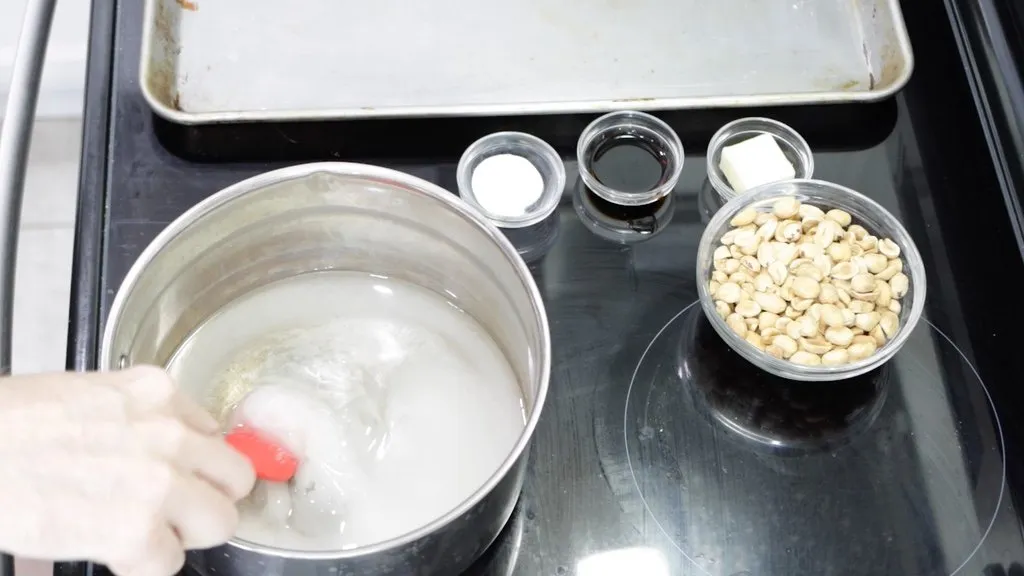 Hand stirring a pot of sugar and corn syrup next to a bowl of peanuts.
