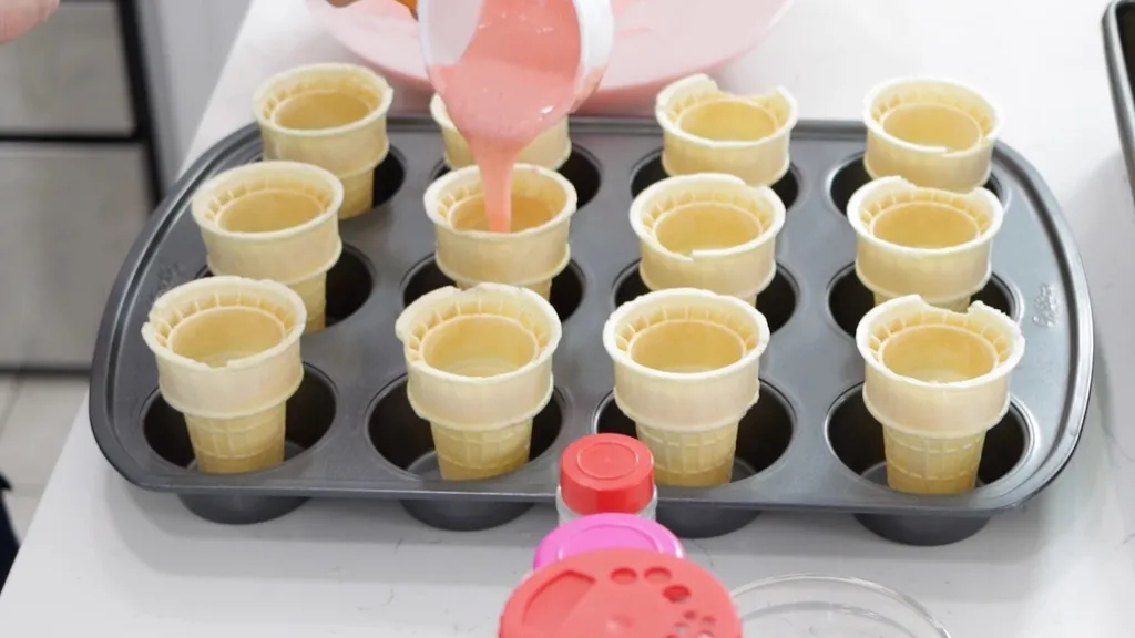 Hand pouring strawberry cake batter into ice cream cones in a muffin pan.