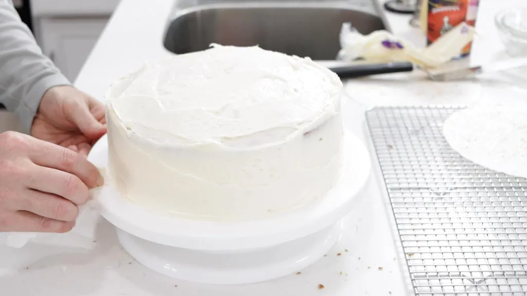 Hand cleaning the edges of a vanilla cake with a damp paper towel.