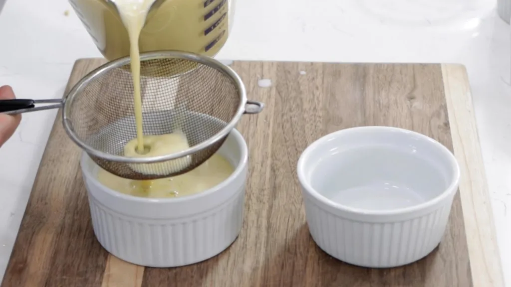 Creme brulee custard being poured through a strainer into white ramekins.