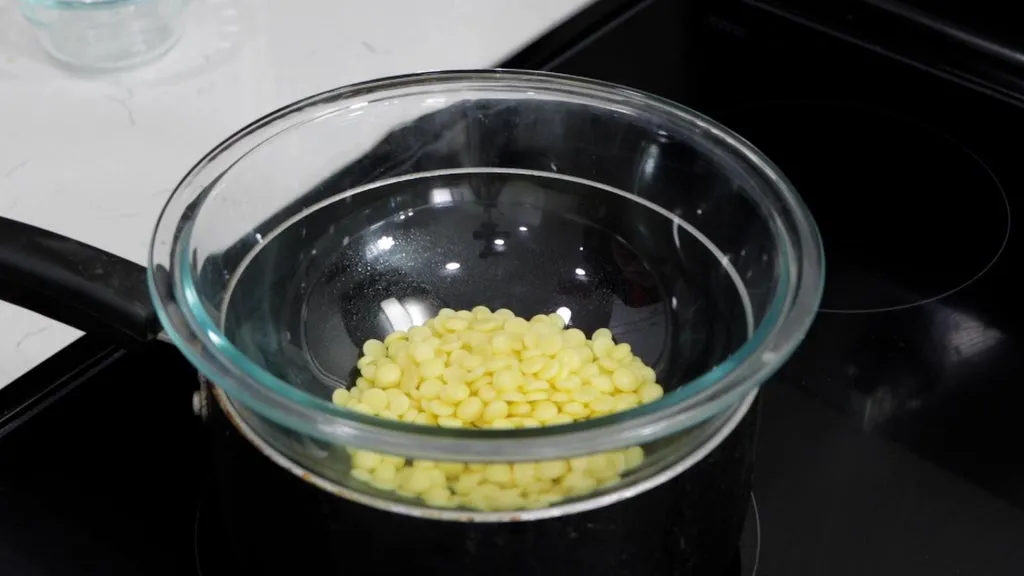 Cocoa butter discs in a pyrex bowl on top of a small pot on a stovetop burner.
