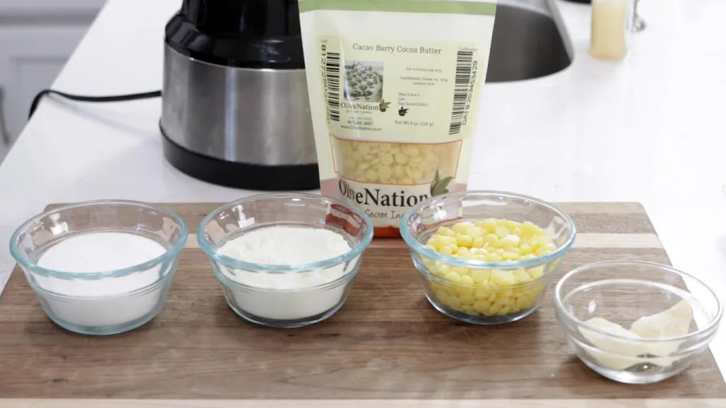 Sugar, milk powder, and cocoa butter in bowls on top of a cutting board.