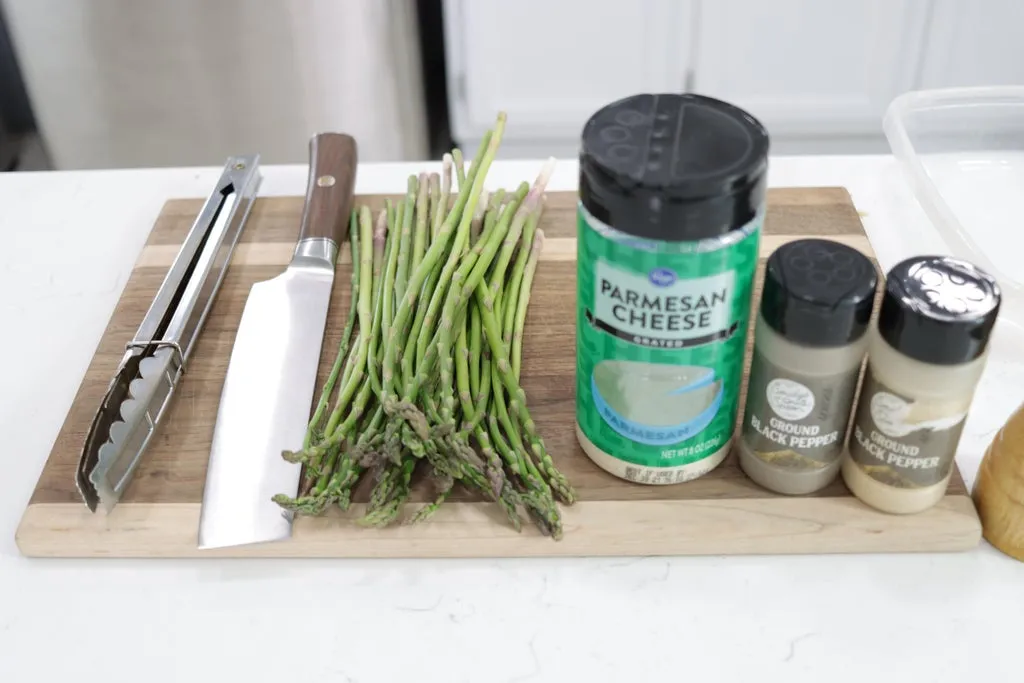 Raw asparagus, knife, tongs, and seasonings on a cutting board.