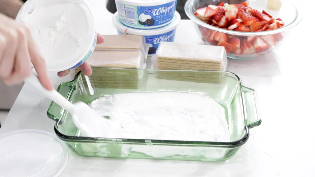 Spatula spreading whipped topping on a the bottom of a green glass baking dish.