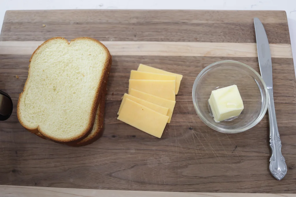 Two slices of bread, several small slices of cheese, and butter in a glass dish all on a cutting board.