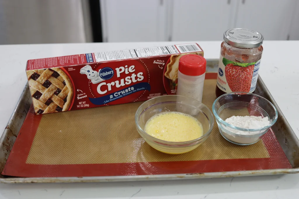 Pie crusts, strawberry preserves, egg wash, and flour on a sheet pan.