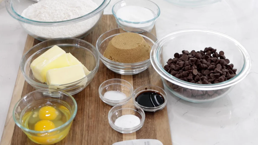 Several ingredients like sugar, butter, chocolate chips, in glass bowls on a wooden cutting board.