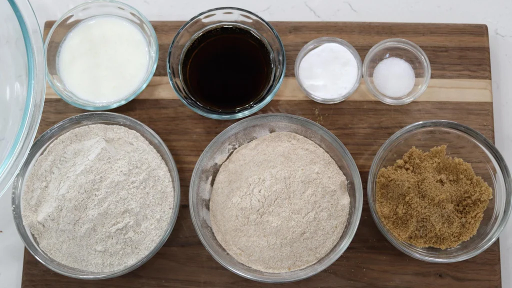 Several bread ingredients in small glass bowls on top of a wooden cutting board.