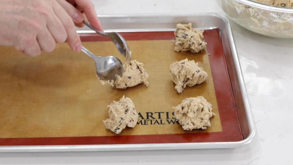 Scooping chew chocolate chip cookies dough onto a sheet pan with a silicone baking mat.
