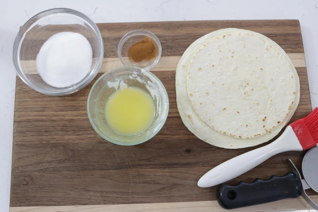 Flour tortillas, melted butter, cinnamon, and sugar on a wooden cutting board.