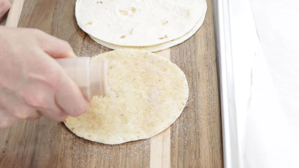Hand shaking cinnamon and sugar on some flour tortillas.