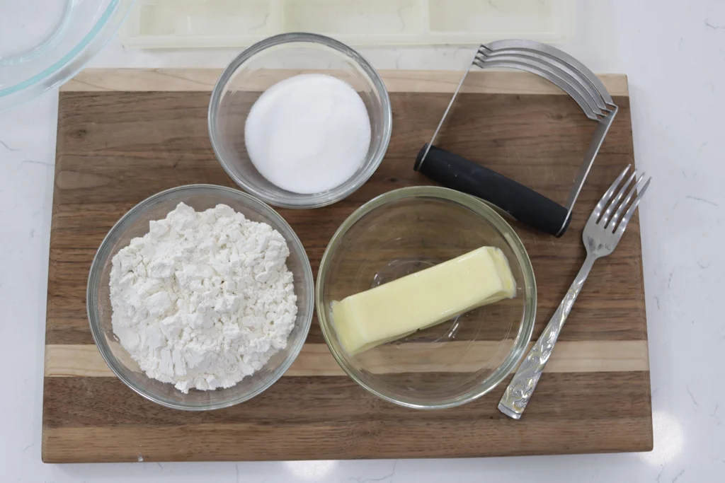 Butter, sugar, and flour in bowls on a wooden cutting board.