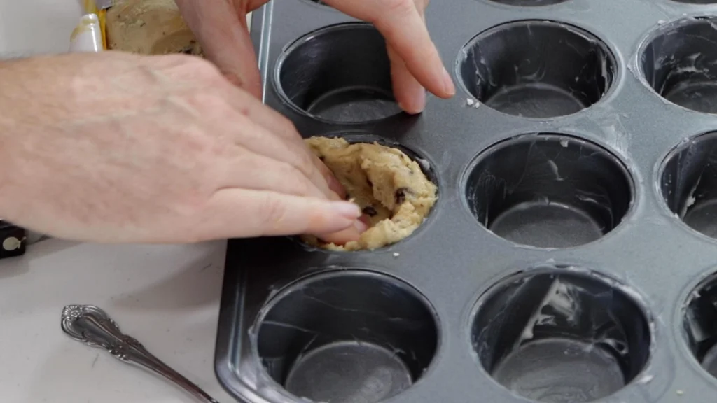 Hand pressing chocolate chip cookie dough into the cavity of a muffin pan.