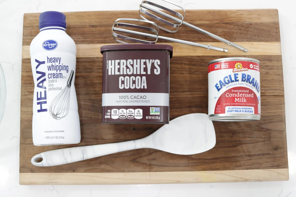 Heavy whipping cream, cocoa powder, and sweetened condensed milk on a wooden cutting board.