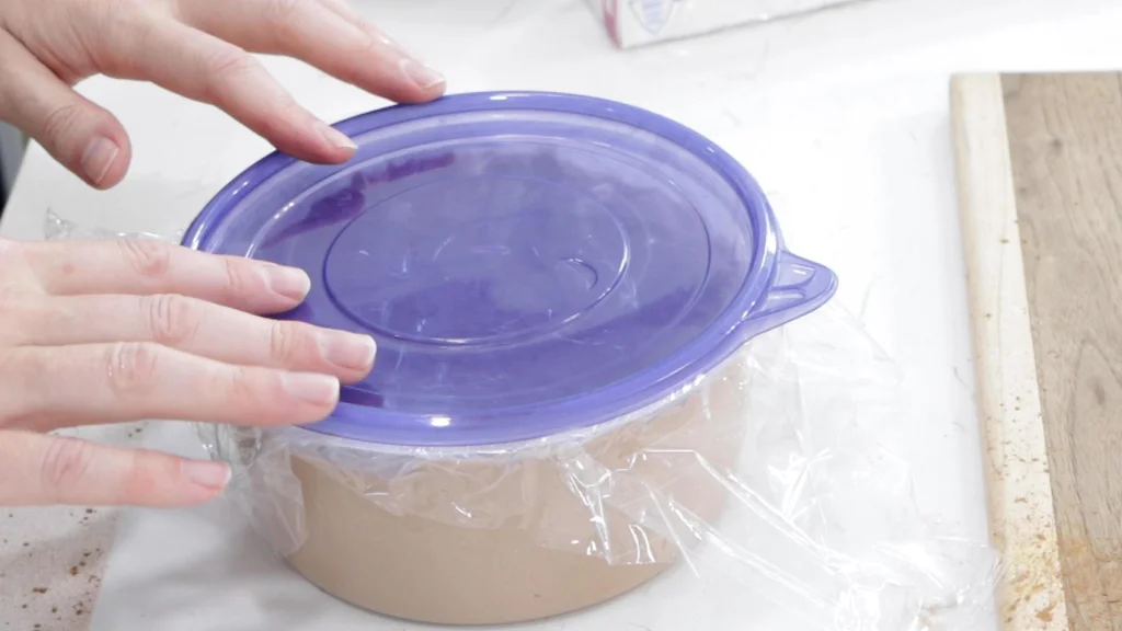 Ice cream base in a plastic container with plastic wrap and a blue lid.