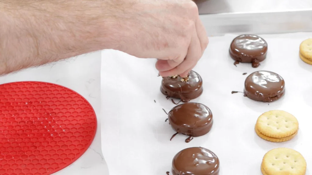 Hand adding chopped nuts to a chocolate covered peanut butter Ritz cracker.