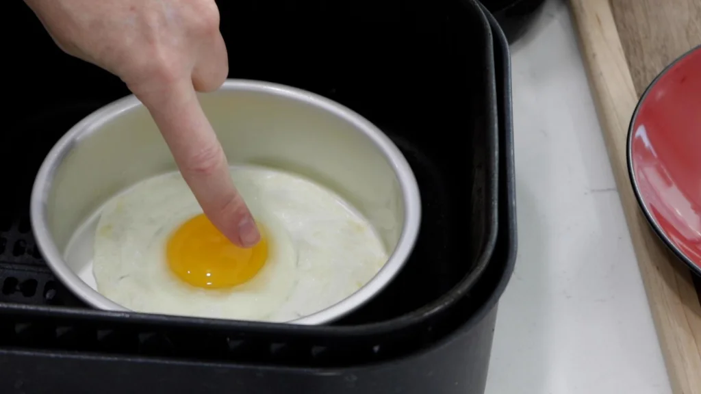 Finger pointing at almost cooked fried egg in a cake pan in air fryer basket.