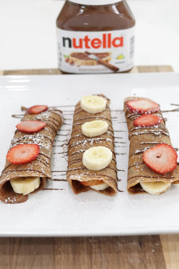 Three Nutella crepes on a white plate with a jar of Nutella behind them.