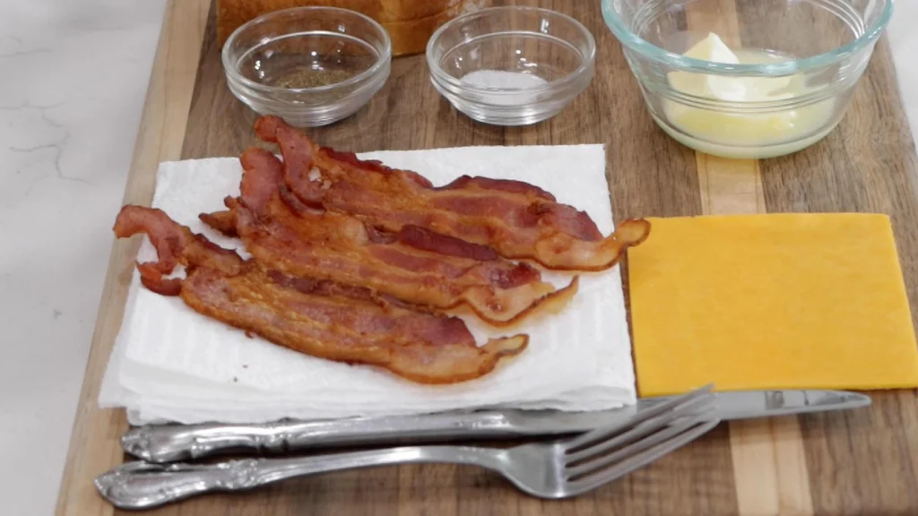 Three slices of cooked bacon next to a slice of cheese on a wooden cutting board.