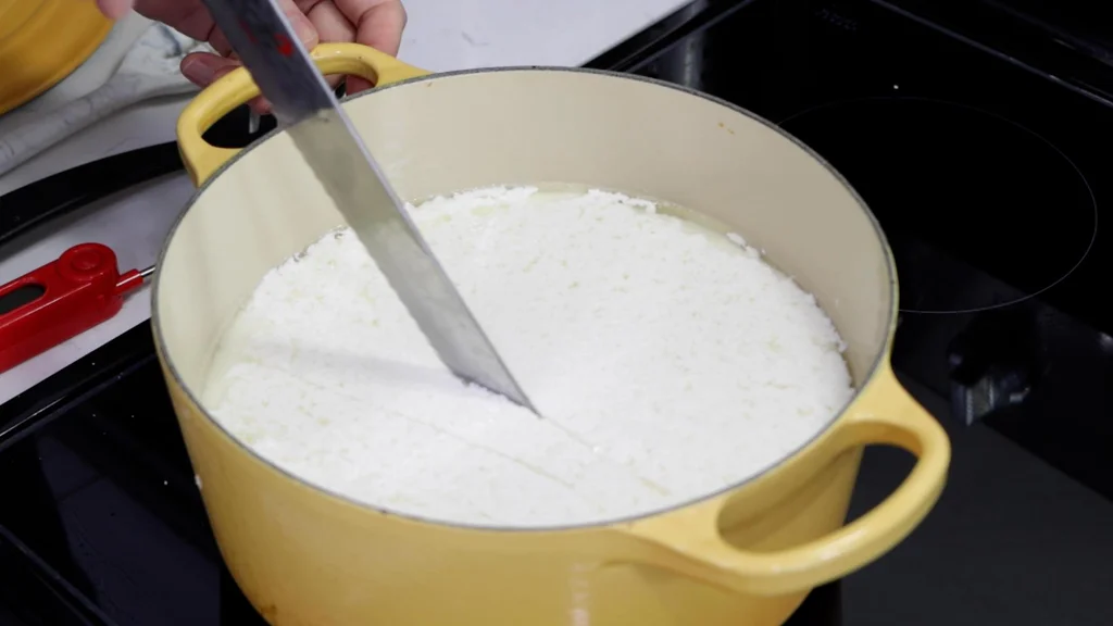 Knife cutting the mozzarella cheese curd in a large yellow pot.