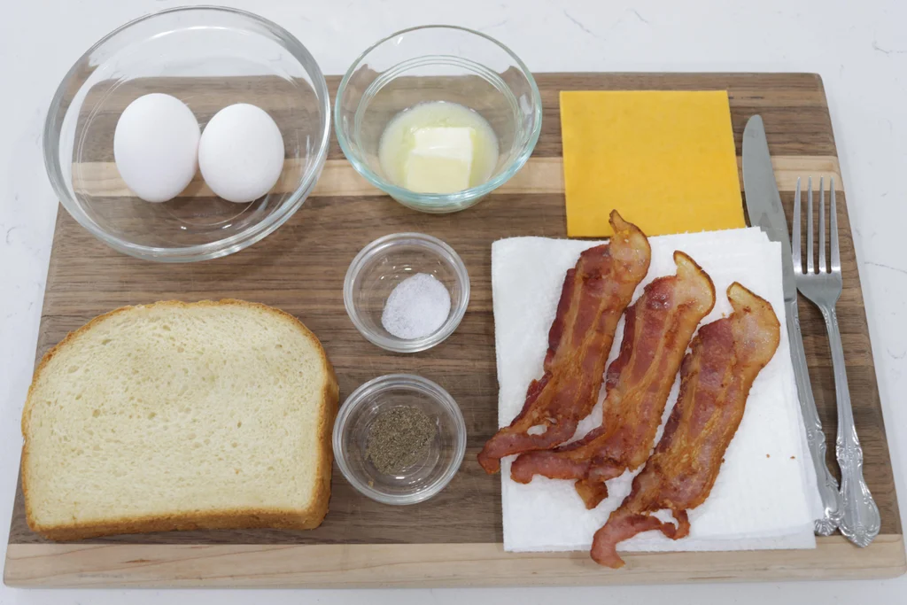 Eggs, bread, butter, cheese, and bacon on a wooden cutting board.