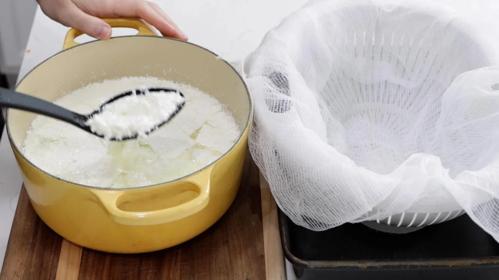 black slotted spoon scooping the mozzarella cheese curds out of the pot of whey.