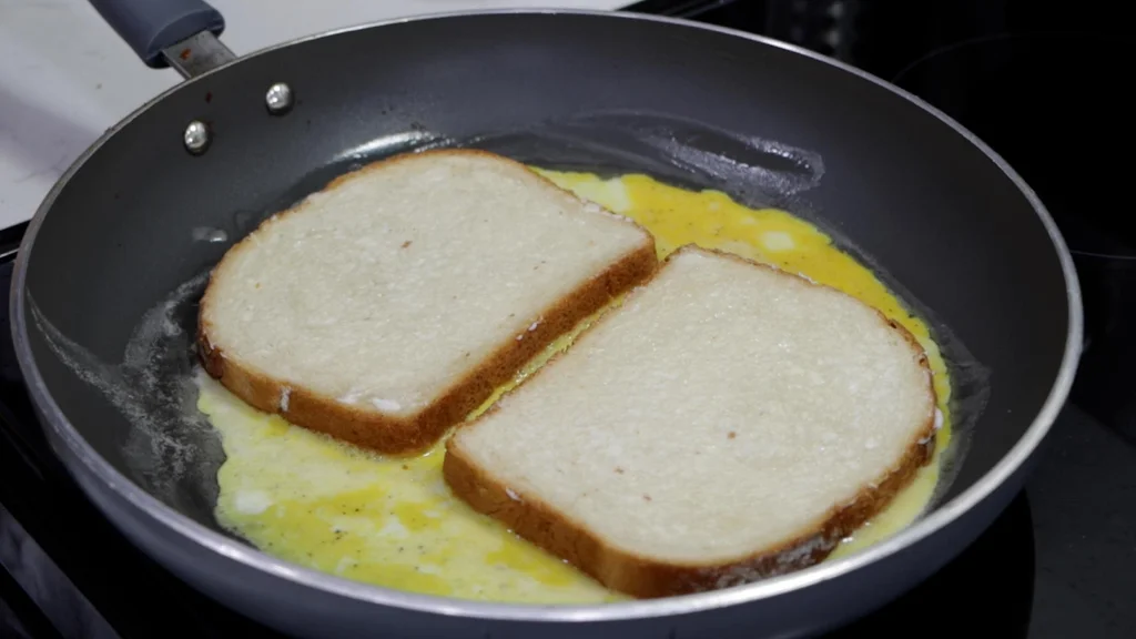 Two butter slices of bread laying on top of egg mixture in a skillet. 