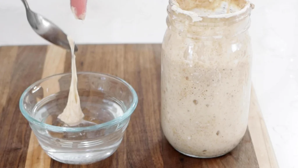 Spoon scooping a teaspoon of sourdough starter into a bowl of water.